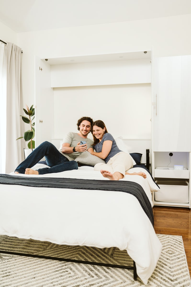 Two people sitting on the Avalon Murphy bed in a compact living space with a white wall and minimal decor.