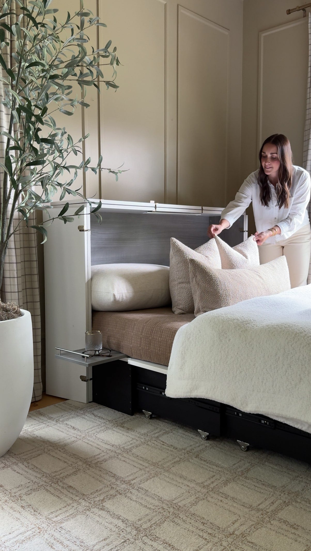 Woman adjusting pillows on a cabinet bed with pull-out drawer in a room with a plant and curtains.