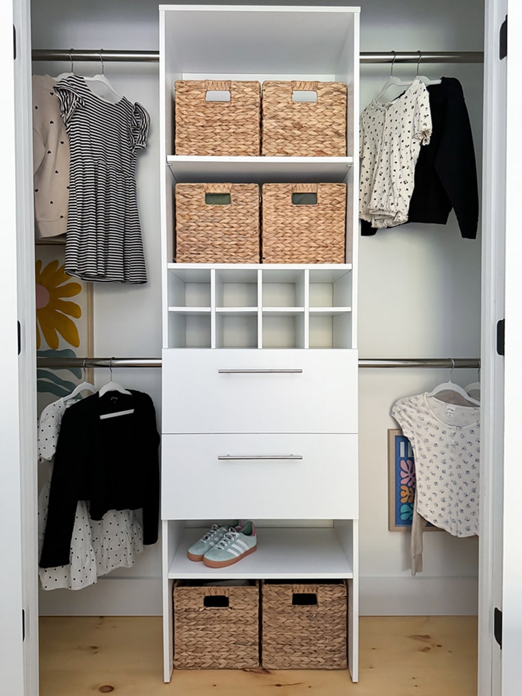 Carina collection closet with white storage unit, wicker baskets, and clothes hanging