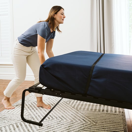 Woman setting up a navy blue mattress on a metal frame in a room with white walls and wooden flooring.
