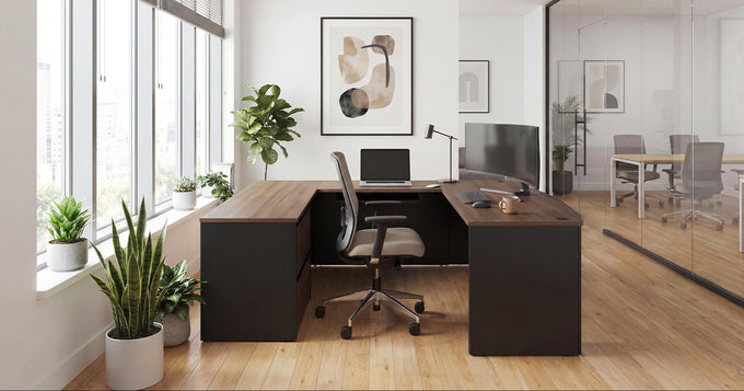 A modern L-shaped home office desk setup in a bright, contemporary room. The desk has a dark brown wood-grain top and a matte black base, paired with a black and gray swivel office chair.