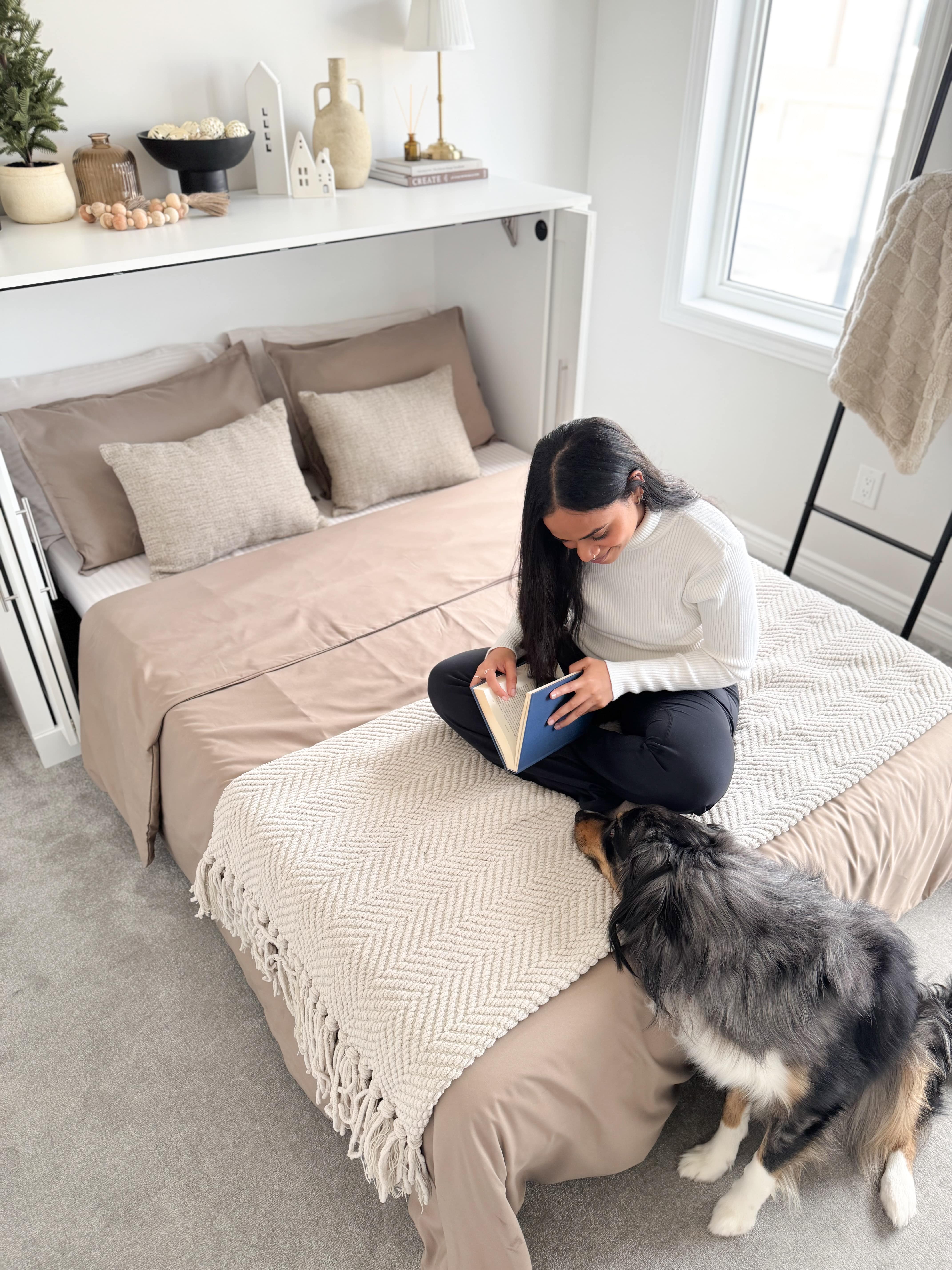 Woman sitting on a cabinet bed with a dog, using a tablet in a bedroom setting.