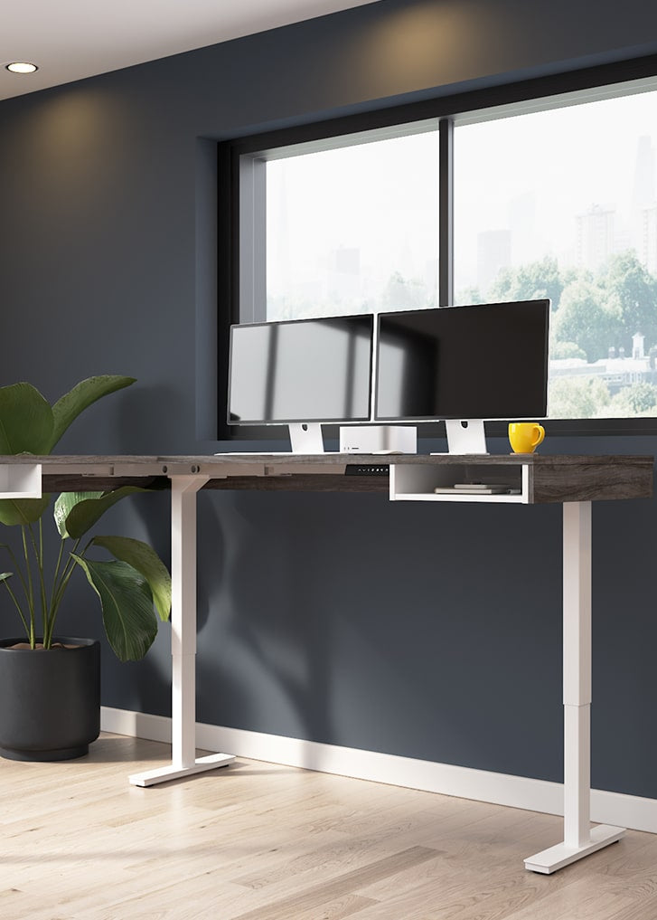 Modern office setup with a standing desk, computer monitors, and a plant in a room with large windows.