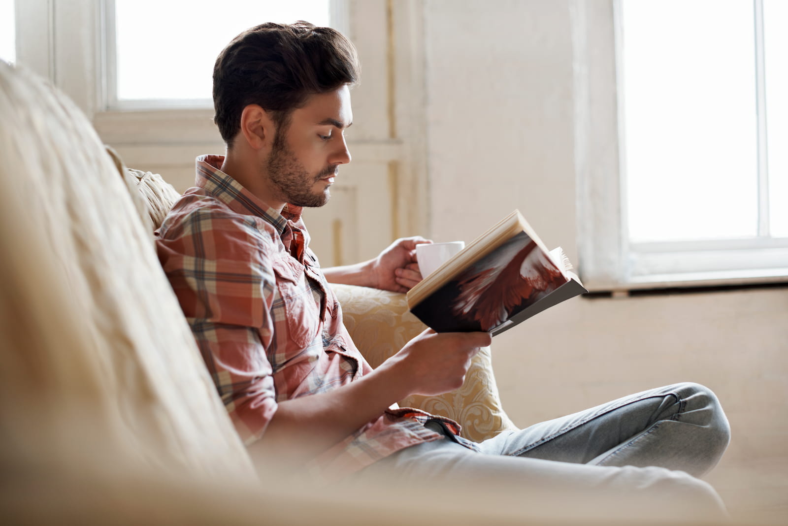 man reading book on the couch with coffee in hand