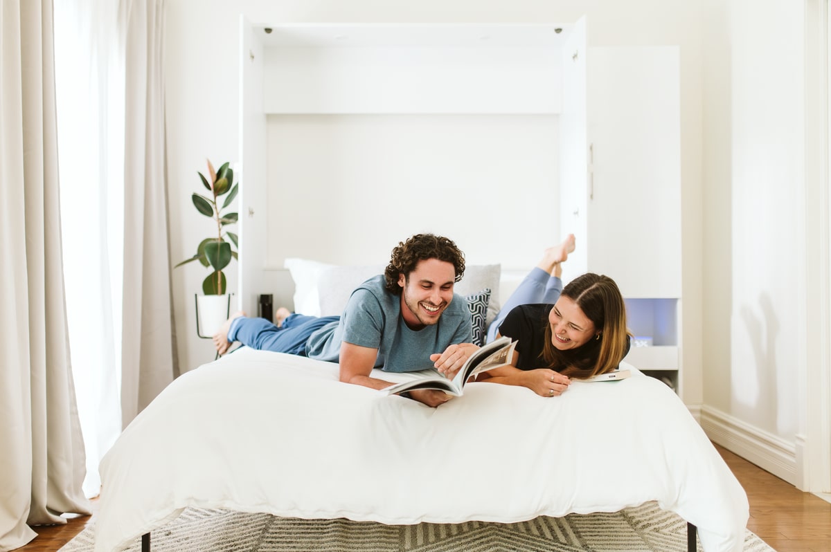 Couple lying on a Bestar Murphy bed, reading together in a bright, minimalist bedroom.